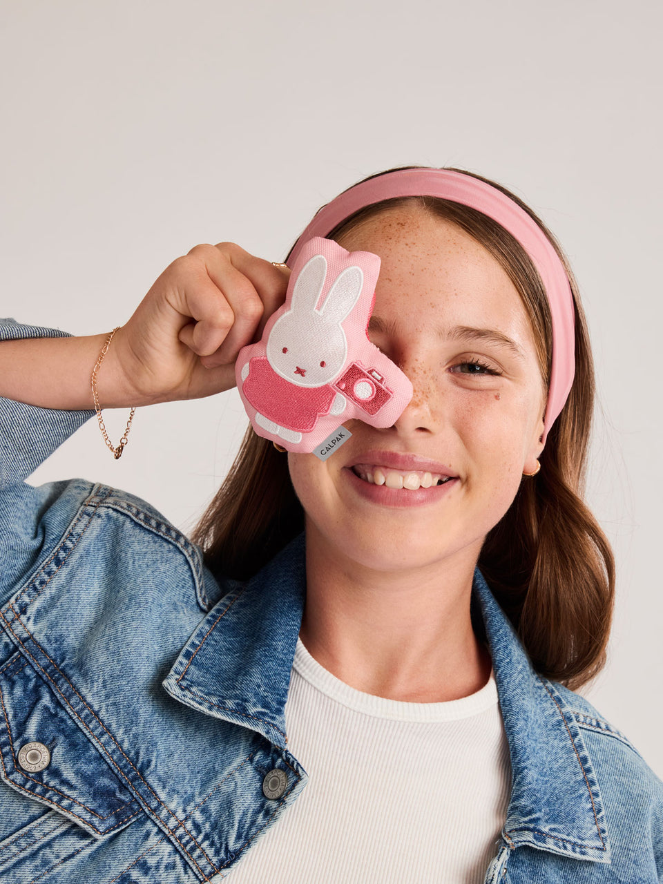 Girl posing with the Miffy x CALPAK Pluffy Bag Charm keychain with a pink border and Miffy holding a camera; APLF2501-MIFFY-MAKES-MEMORIES