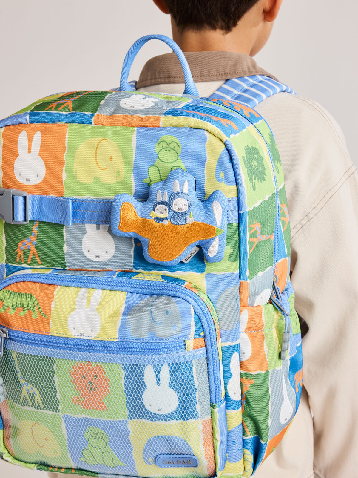 Child wearing Miffy x CALPAK Pluffy Bag Charm keychain with a blue border and Miffys holding a carrot on an orange airplane attached to a color Kids CALPAK Kids Backpack; APLF2501-MIFFY-ON-THE-WAY