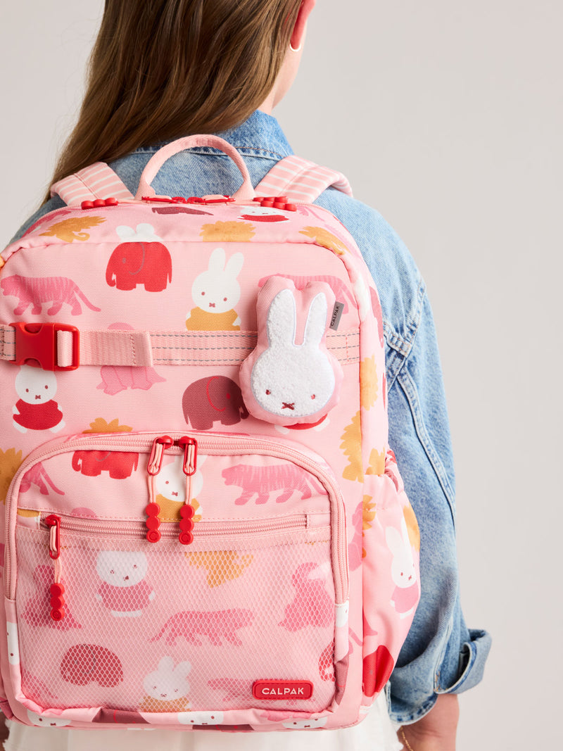 Plush Miffy x CALPAK Pluffy Bag Charm keychain with a pink border and Miffy being worn by a child with a colorful Kids CALPAK Kids Backpack