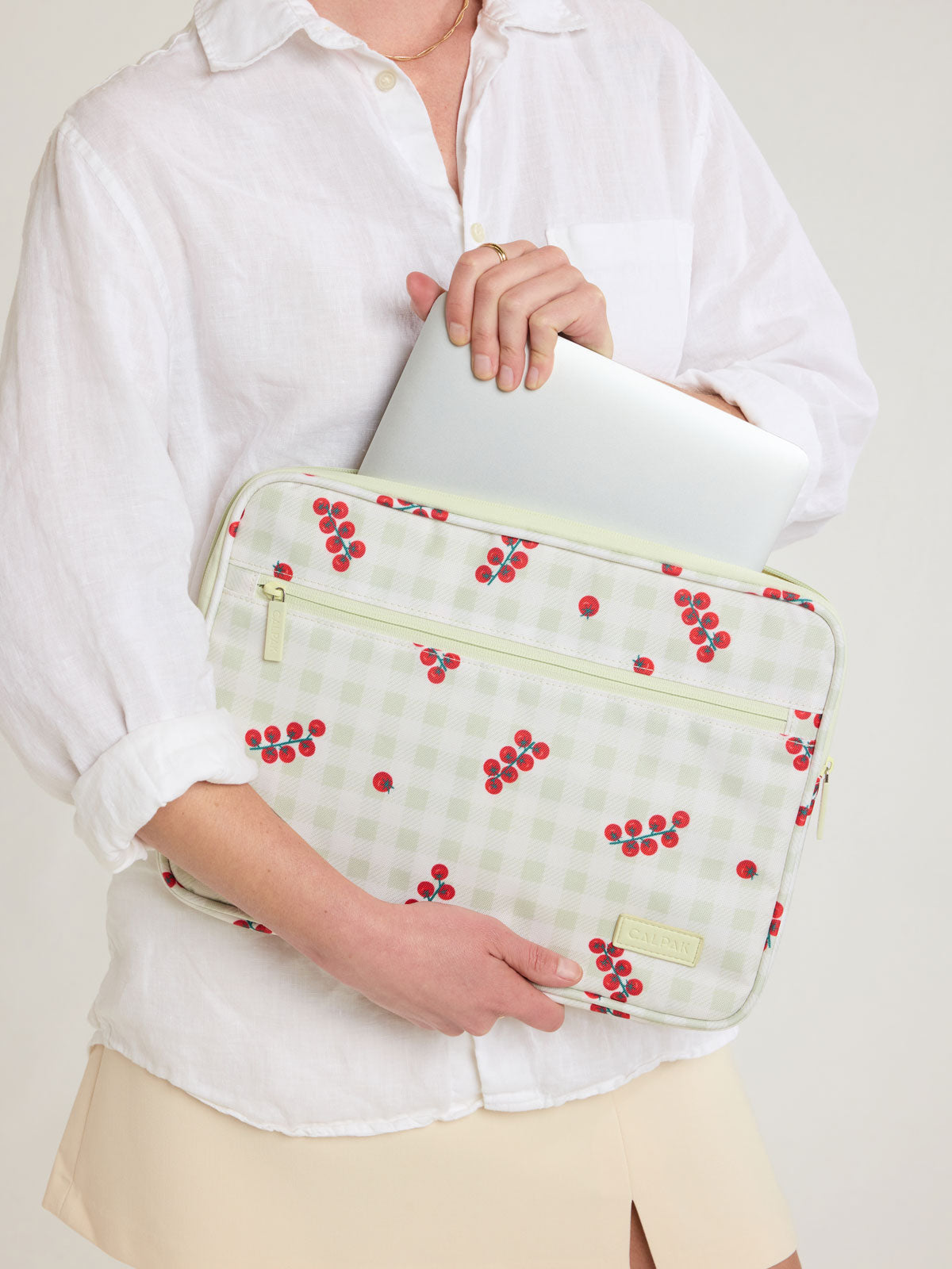 Person putting laptop into CALPAK 13"-14" Padded Laptop Sleeve in white and green gingham pattern with tomato print; ALP2213-CELADON-TOMATO-PICNIC