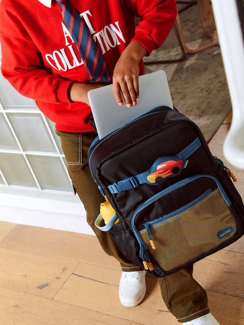 CALPAK Kids Large Backpack in Black for ages 8-12 with child putting in notebook into Large Backpack with Car Pluffy Bag Charm