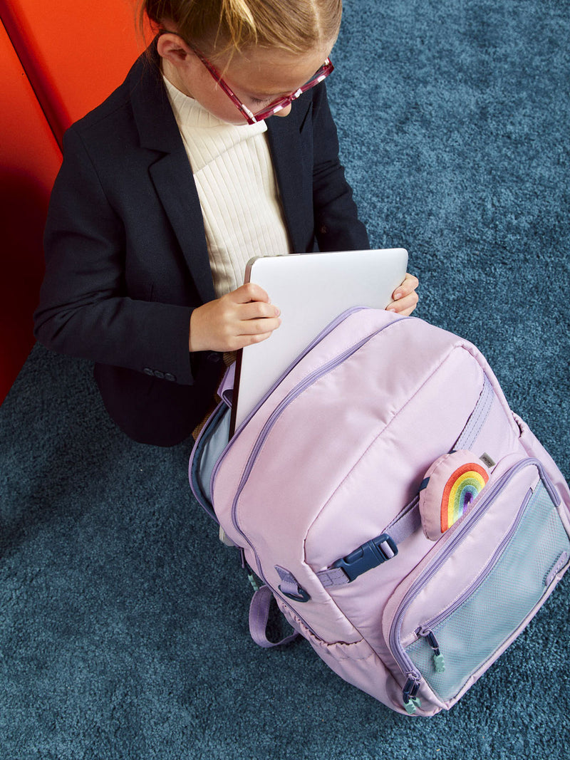 CALPAK Kids Large Backpack in pink lavender Orchid Pink for ages 8-12 with child putting in laptop into Large Backpack with Rainbow Pluffy Bag Charm