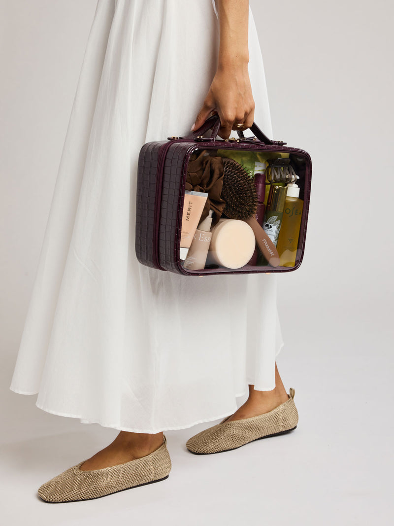 A person in a flowing white dress holds  a CALPAK Large Clear Cosmetics Case in deep red Dark Cherry against a white background