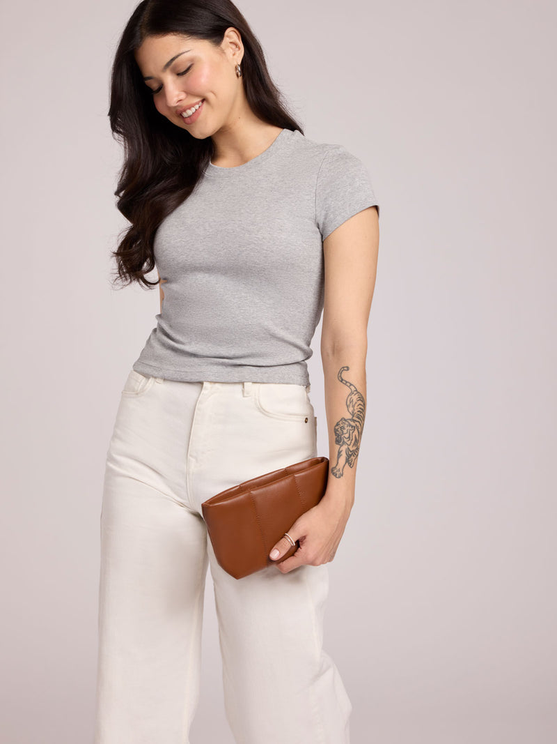 A woman wearing a light gray short-sleeve crewneck t-shirt and off-white wide-leg jeans, holding a CALPAK Luka 1 Quart Pouch in Honey Brown light gray studio background