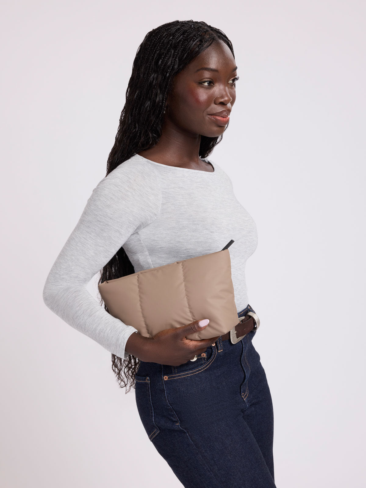 A woman wearing a light gray long-sleeve top and dark blue jeans holds a beige quilted puffer clutch bag against a plain light gray studio background; ZLPM2601-CHOCOLATE