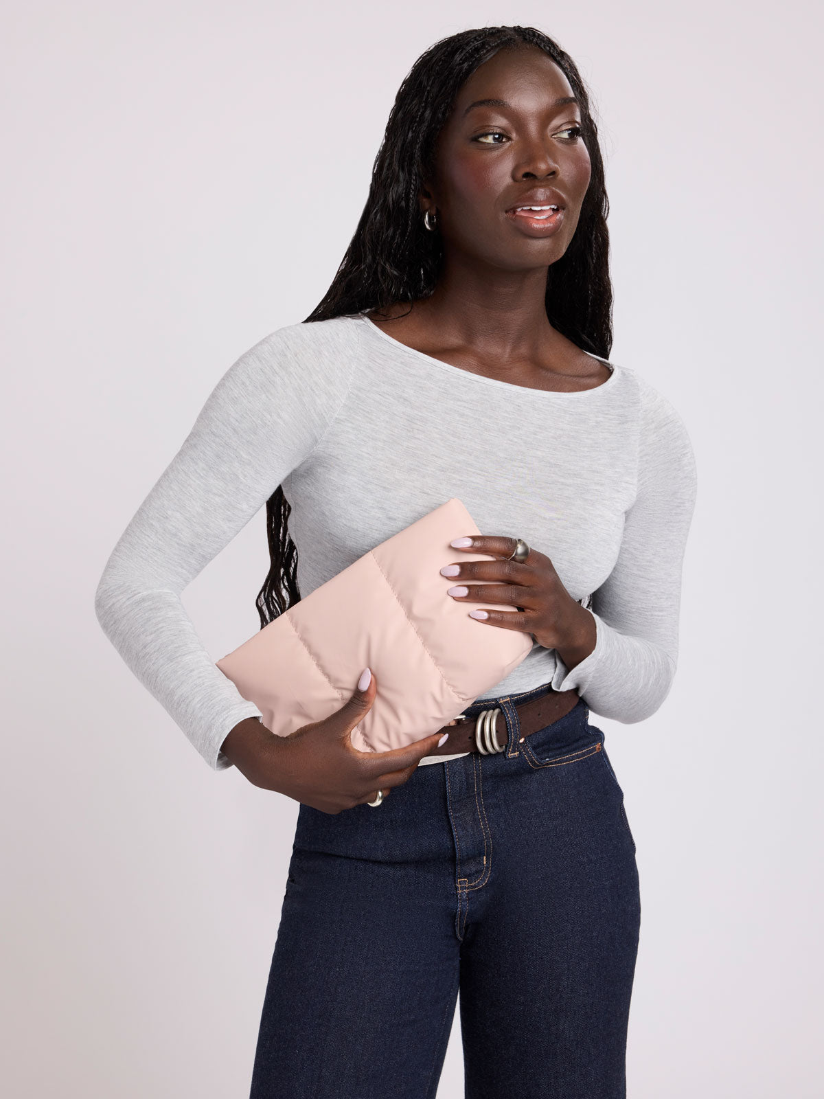 A woman modeling a light gray long-sleeve top and dark blue jeans, holding a light pink puffer clutch bag against a plain light gray background; ZLPM2601-ROSE-QUARTZ
