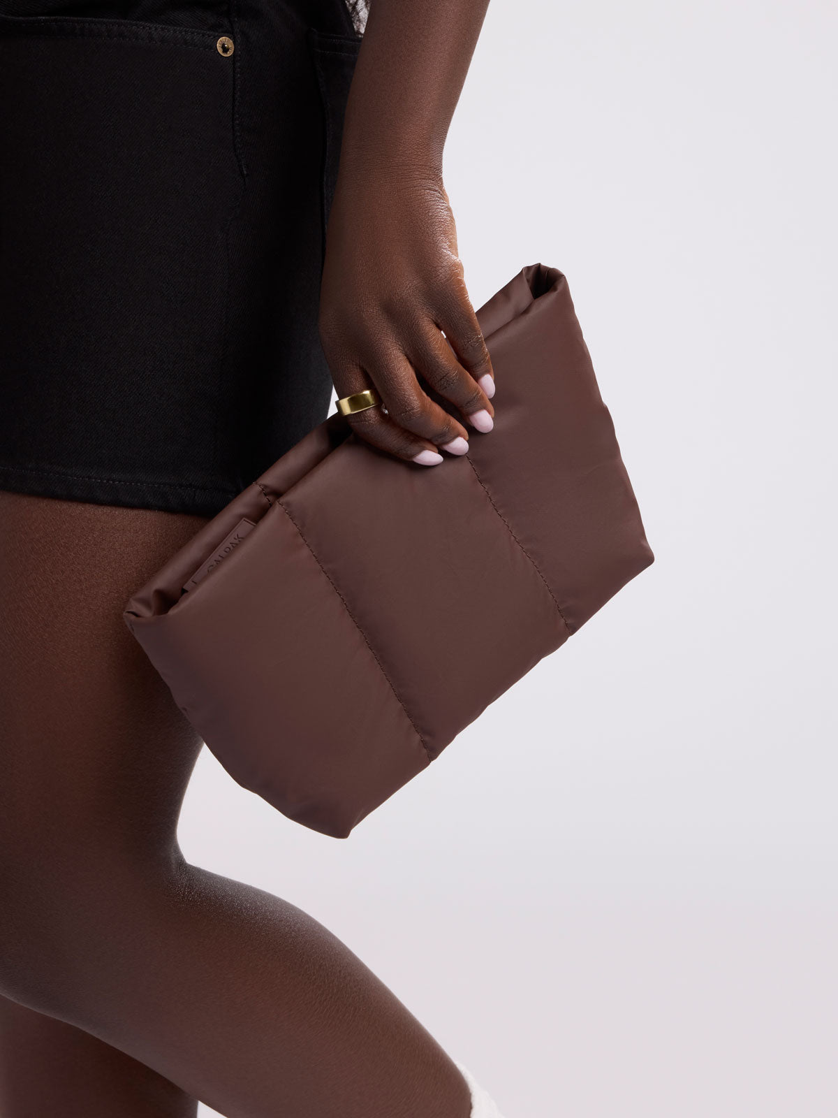 A close-up of a person holding a brown puffer-style clutch bag against a light gray studio background; ZLPM2601-WALNUT