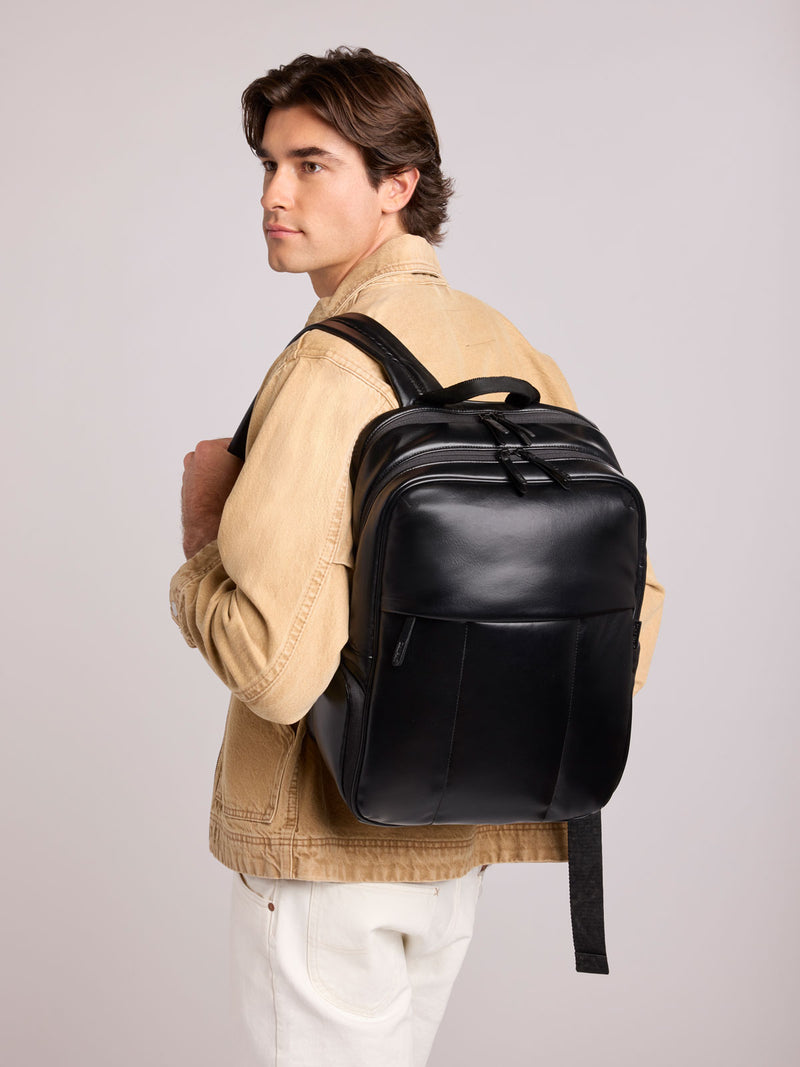 A man wearing a beige jacket and white pants is shown from the back and side, carrying a CALPAK Luka 15in Laptop Backpack in Black Vegan Leather against a light gray studio background