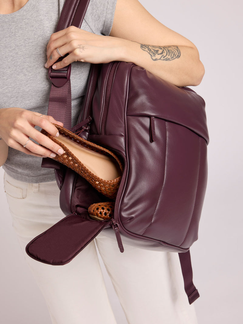 A person wearing a gray sleeveless top and white pants carries a CALPAK Luka 15in Luka Laptop Backpack in Dark Cherry Vegan Leather while putting in shoes in the bottom shoe compartment