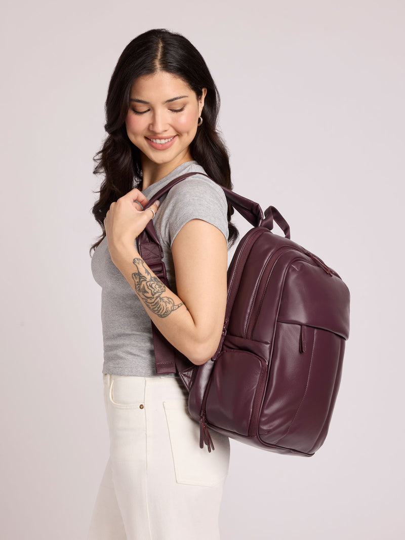 A woman with long dark hair, wearing a gray t-shirt and white pants, carries a CALPAK Luka 15in Laptop Backpack in Dark Cherry Vegan Leather over her shoulder against a light gray studio background