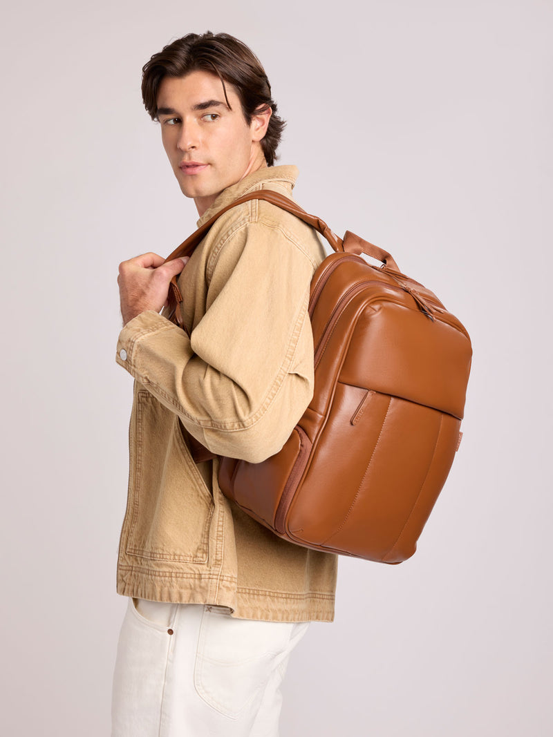 A man wearing a tan denim jacket and white pants carries a CALPAK Luka 15in Laptop Backpack in Honey Brown Vegan Leather over his shoulder, photographed against a light gray studio background