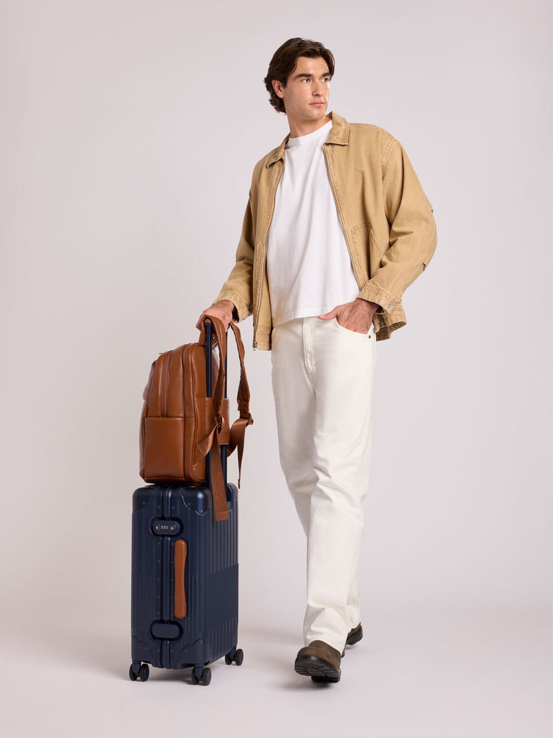 A man stands in a studio wearing a beige overshirt, white T-shirt, and white trousers, holding a CALPAK Luka 15in Laptop Backpack in Honey Brown Vegan Leather on top of a navy Vault Carry On suitcase
