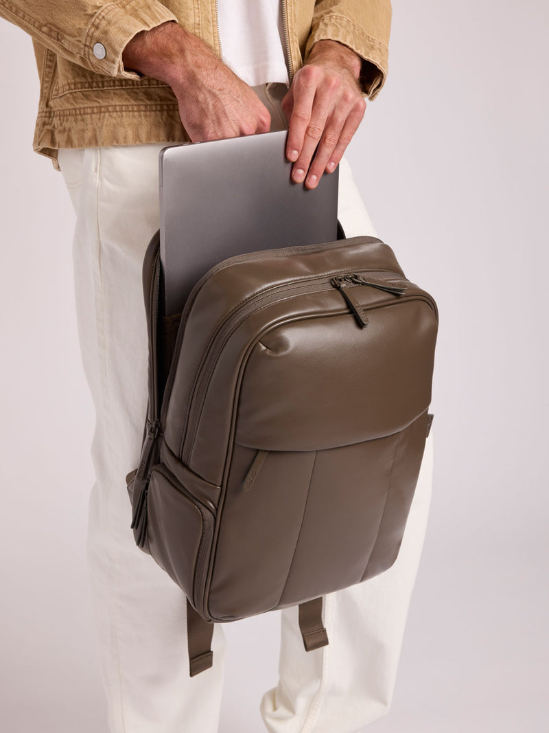 A person wearing a tan jacket and white pants holds a CALPAK Luka 15in Laptop Backpack in Mushroom brown in Vegan Leather with a laptop tucked into the front pocket, against a plain light gray studio background