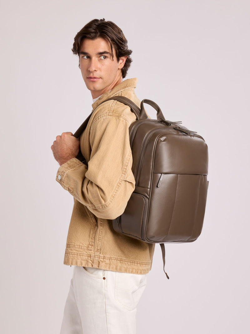 A man wearing a tan jacket and white pants carries a CALPAK Luka 15in Laptop Backpack in Mushroom brown Vegan Leather on his shoulder, photographed against a light gray studio background