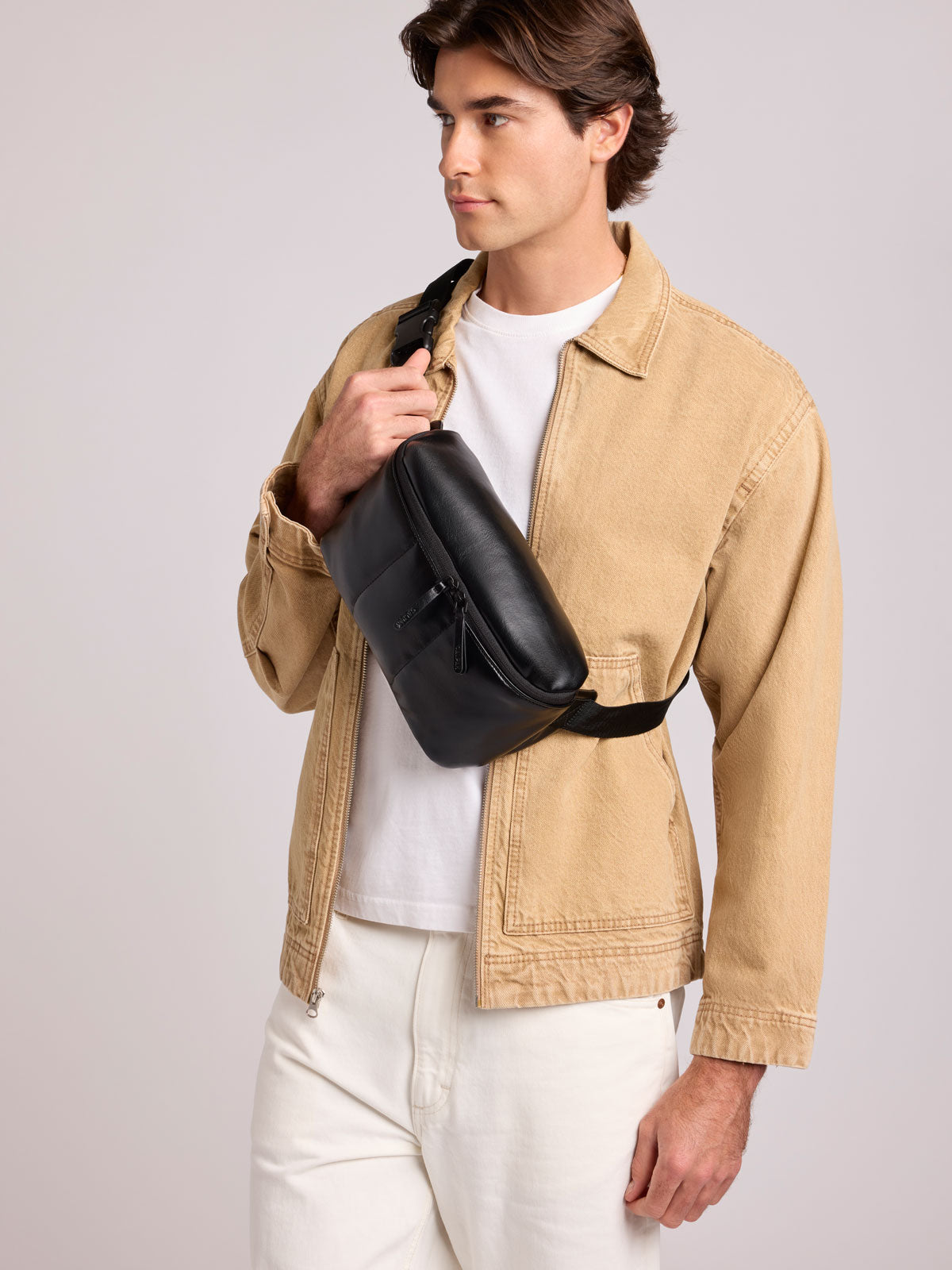 A man wearing a beige overshirt, white t-shirt, and white trousers models a CALPAK Luka Belt Bag in Black Vegan Leather;  BB1901-BLACK-PU