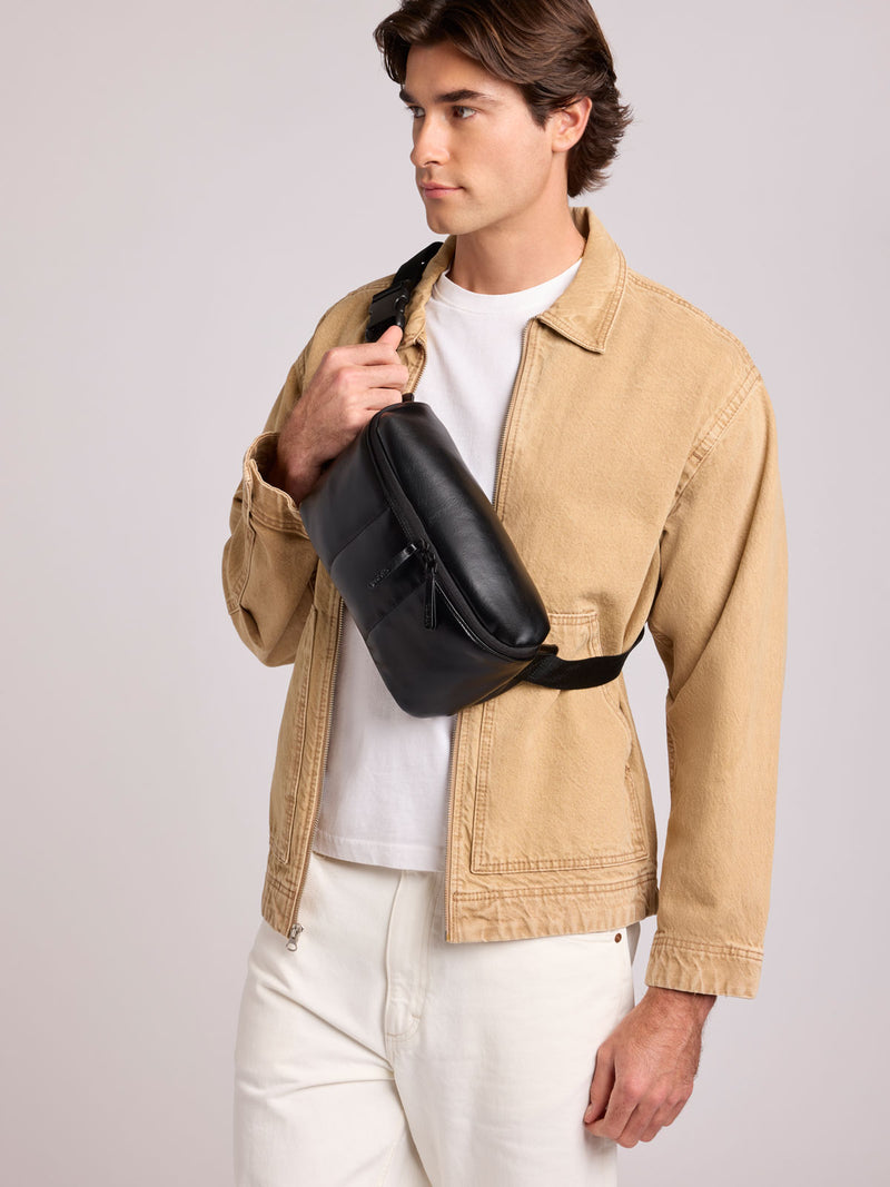 A man wearing a beige overshirt, white t-shirt, and white trousers models a CALPAK Luka Belt Bag in Black Vegan Leather
