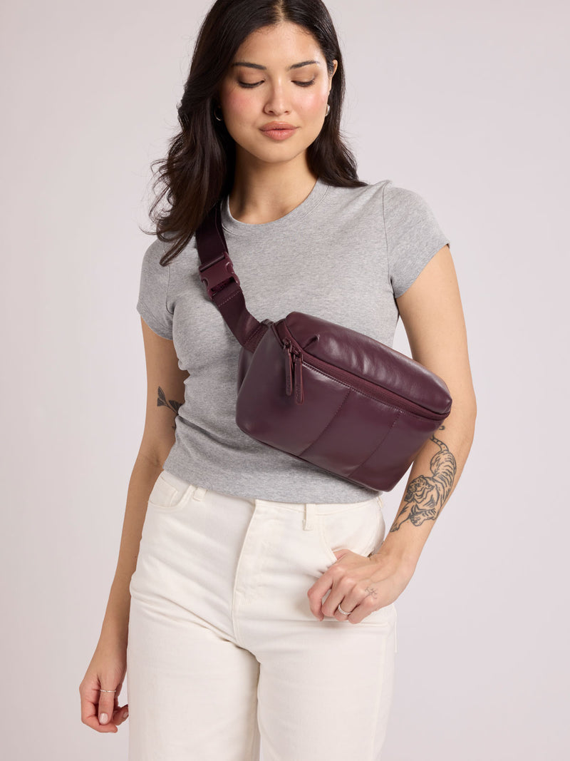 A woman wearing a heather gray t-shirt and white pants models a CALPAK Luka Belt Bag in Dark Cherry Vegan Leather  against a light gray studio background