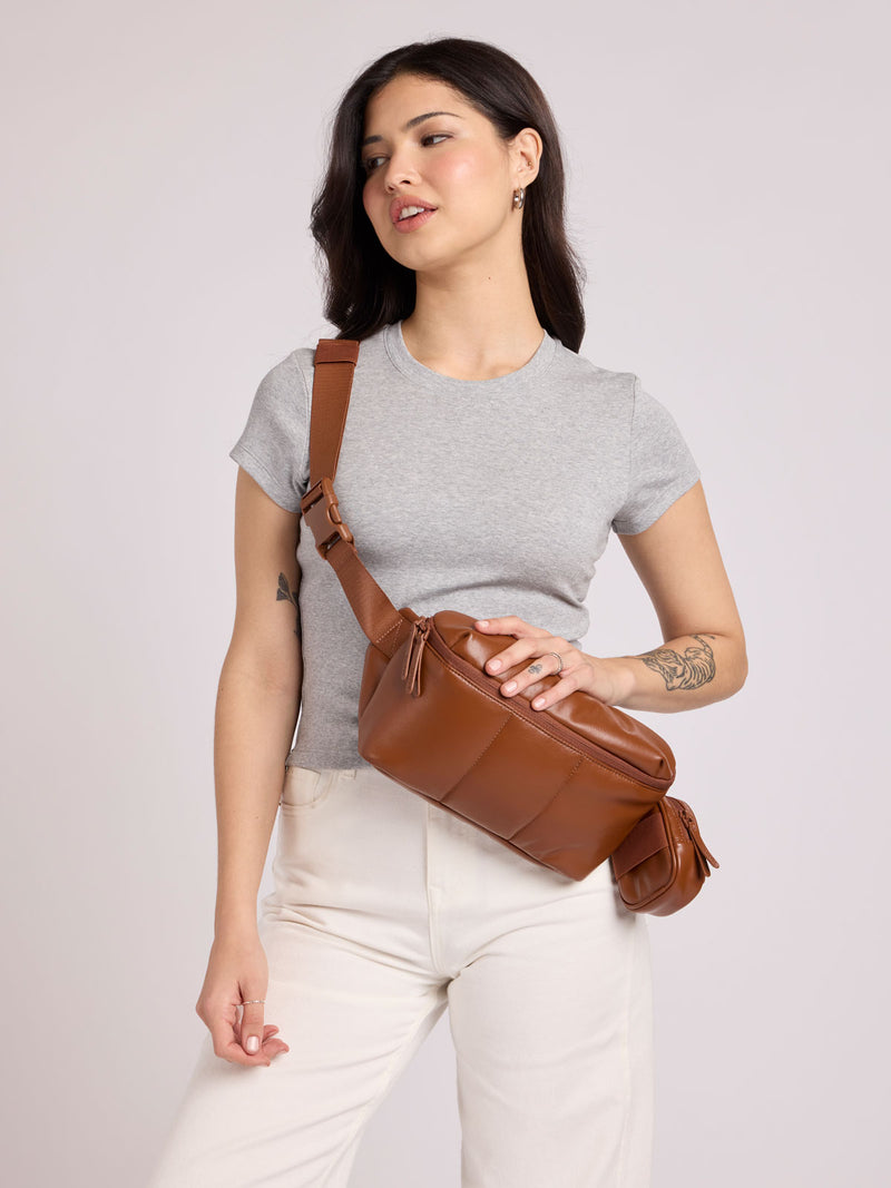 A woman wearing a light gray t-shirt and white pants models a CALPAK Luka Belt Bag in Honey Brown Vegan Leather, set against a solid light gray studio background