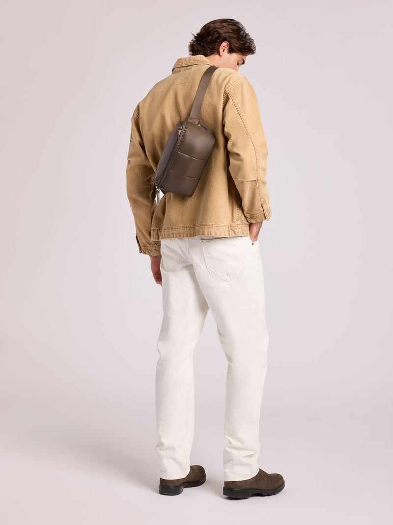 A man is shown from behind wearing a tan canvas utility jacket, white trousers, and brown shoes, with a CALPAK Luka Belt Bag in Mushroom brown Vegan Leather slung over his shoulder, set against a plain light gray studio background