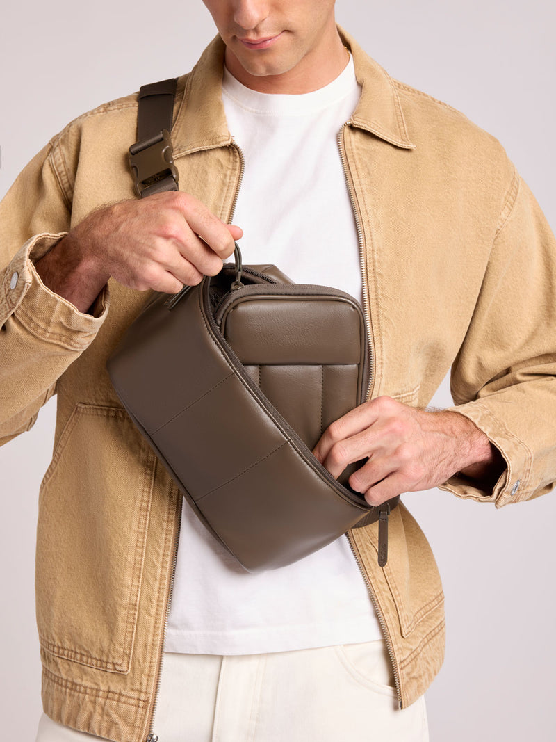 A man wearing a tan overshirt and white t-shirt models a CALPAK Luka Belt Bag in Mushroom Brown Vegan Leather with an adjustable strap, shown from the chest down against a light gray studio background