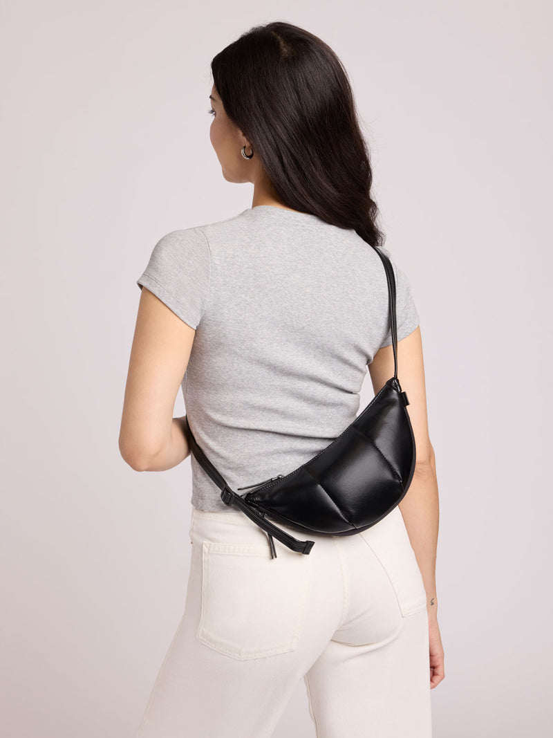 A woman shown from the back wearing a light gray t-shirt and white pants, carrying a CALPAK Luka Crescent Bag in Black Vegan Leather across her back
