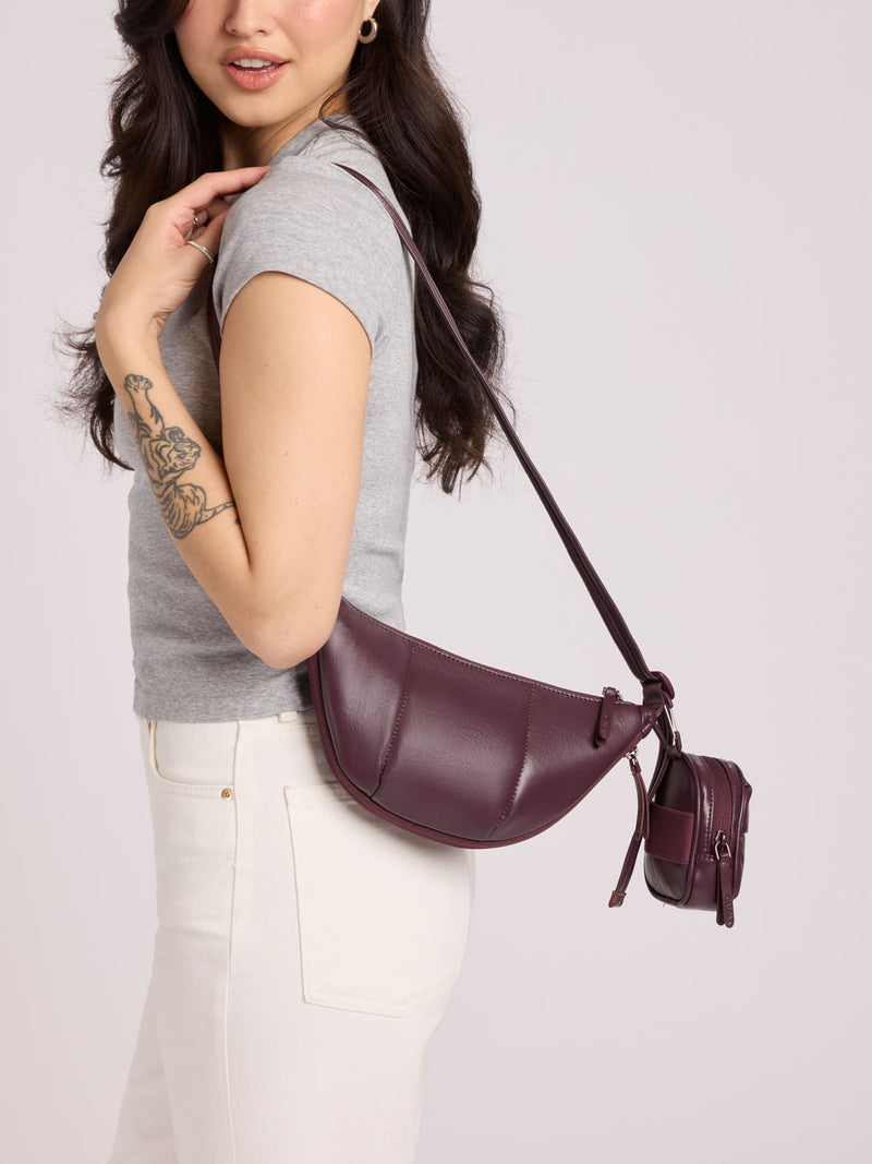 A woman wearing a gray t-shirt and white pants carries a Luka Crescent Bag with the Luka Backpack Zip Bag Charm attached in Dark Cherry Vegan Leather against a light gray studio background