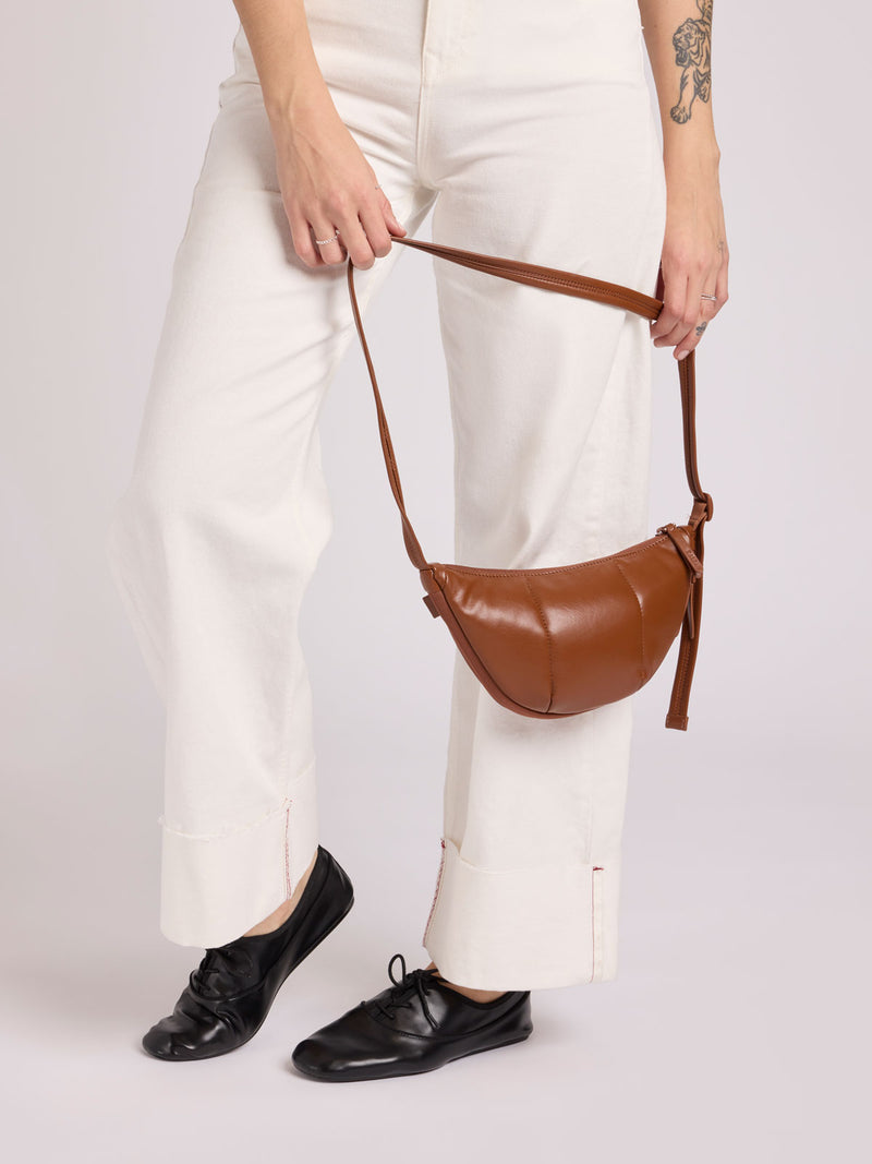 A person wearing white wide-leg trousers and black loafers holds a CALPAK Luka Crescent Bag in Honey Brown Vegan Leather with a long strap against a light gray studio background