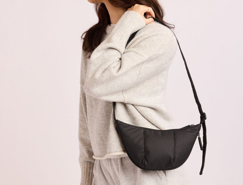 Person wearing a gray sweater with a black CALPAK Luka Crescent Bag on a light pink background