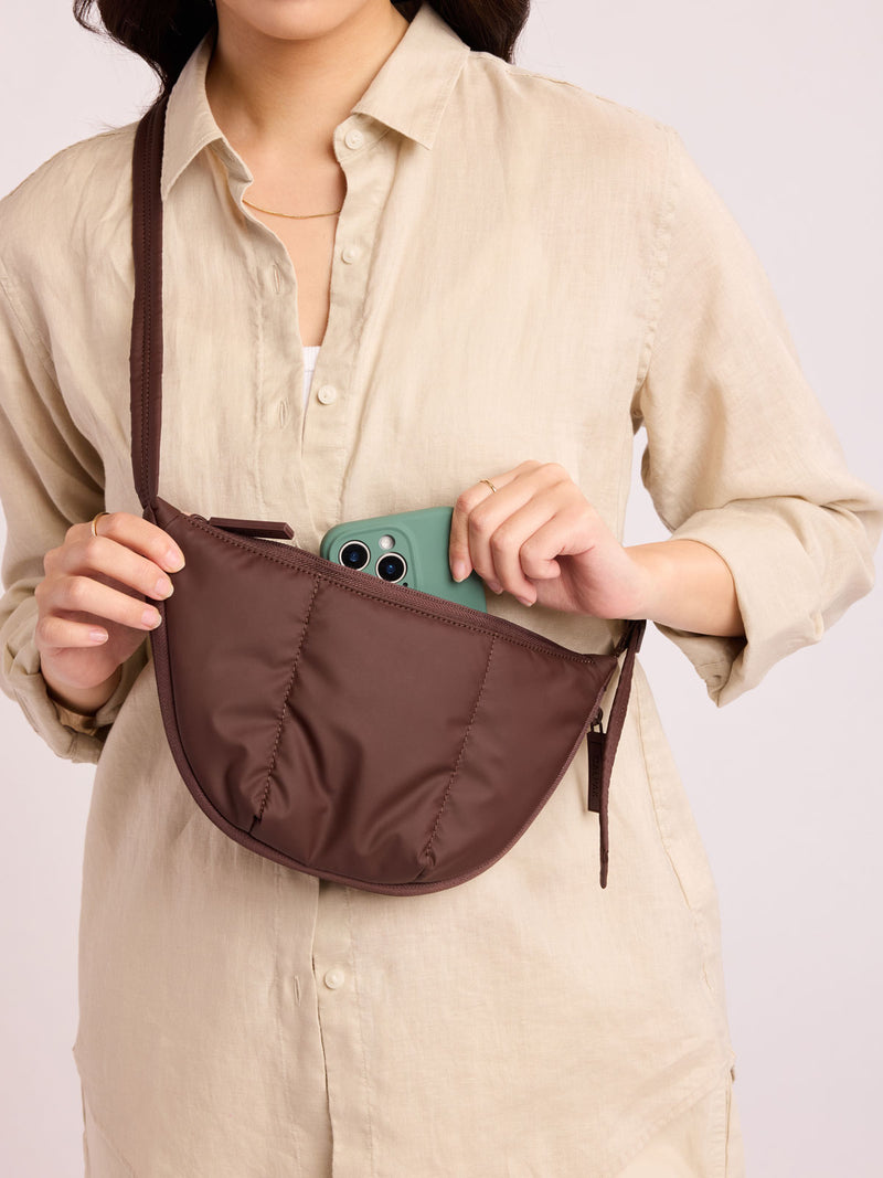 Person with beige outfit putting green phone into CALPAK Luka Crescent Tote bag in brown Walnut with shoulder strap and bag is expandable up to 2" on a white background