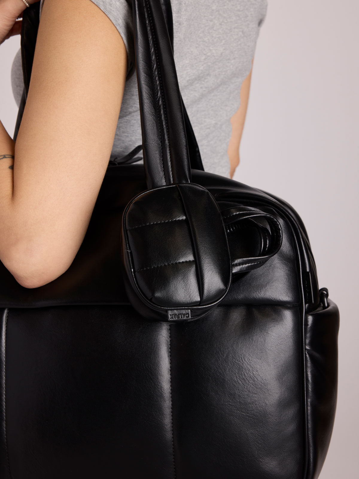 A close-up of a person carrying a CALPAK Luka Duffel in Vegan Leather with a matching CALPAK Luka Duffel Zip Bag Charm attached to the shoulder strap; ALK2501-BLACK-PU