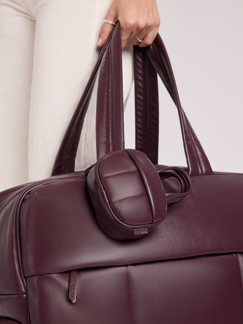 A close-up of a person in white pants holding a CALPAK Luka Duffel in Dark Cherry Vegan Leather with a matching small CALPAK Luka Duffel Zip Bag Charm in Dark Cherry Vegan Leather attached
