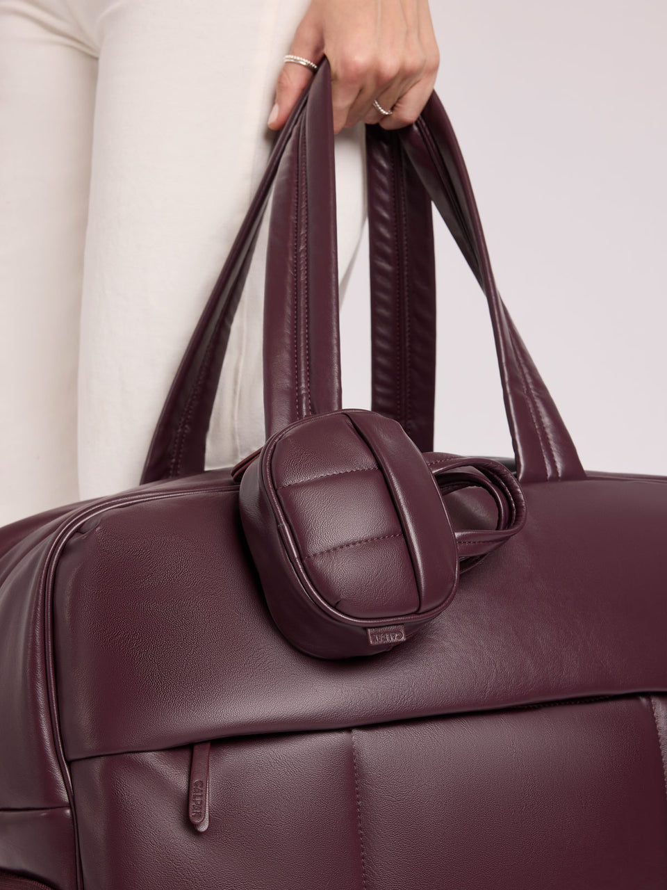 A close-up of a person in white pants holding a CALPAK Luka Duffel in Dark Cherry Vegan Leather with a matching small CALPAK Luka Duffel Zip Bag Charm in Dark Cherry Vegan Leather attached; ALK2501-DARK-CHERRY-PU