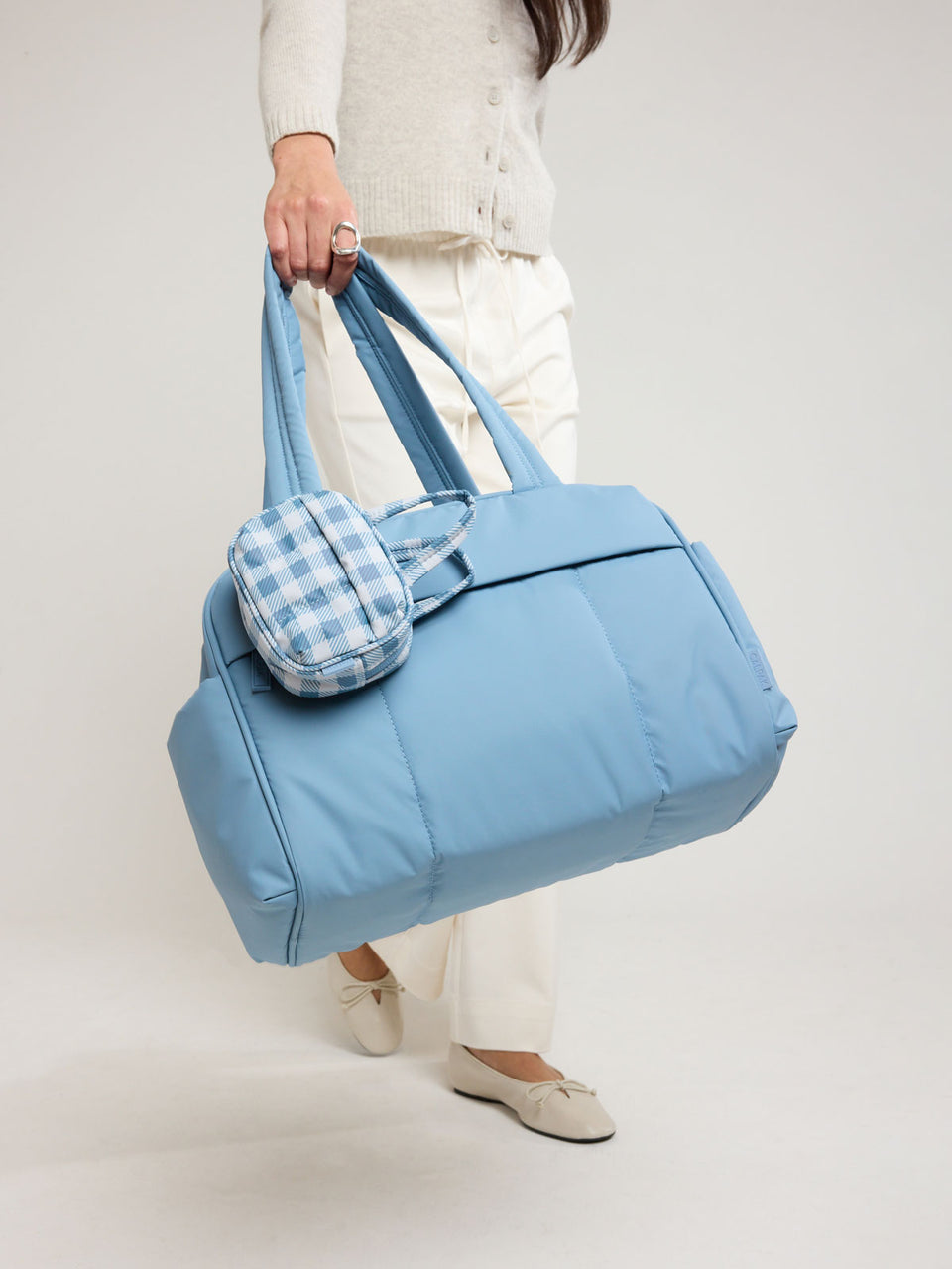 Model with CALPAK Luka Duffel Zip Bag Charm in white and blue gingham print in French Blue Gingham clipped on a bag; ALK2501-FRENCH-BLUE-GINGHAM