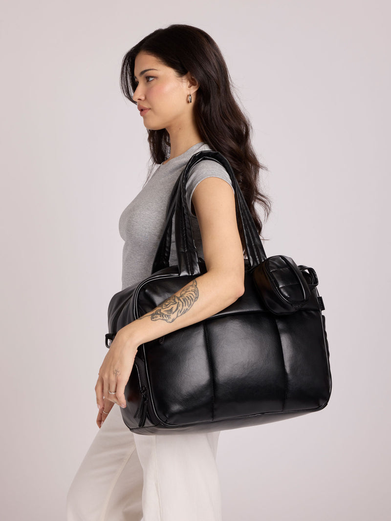 A woman in a gray sleeveless top and white pants carries a Black CALPAK Luka Duffel in vegan leather with dual top handles and a front zippered pocket, photographed against a clean white background