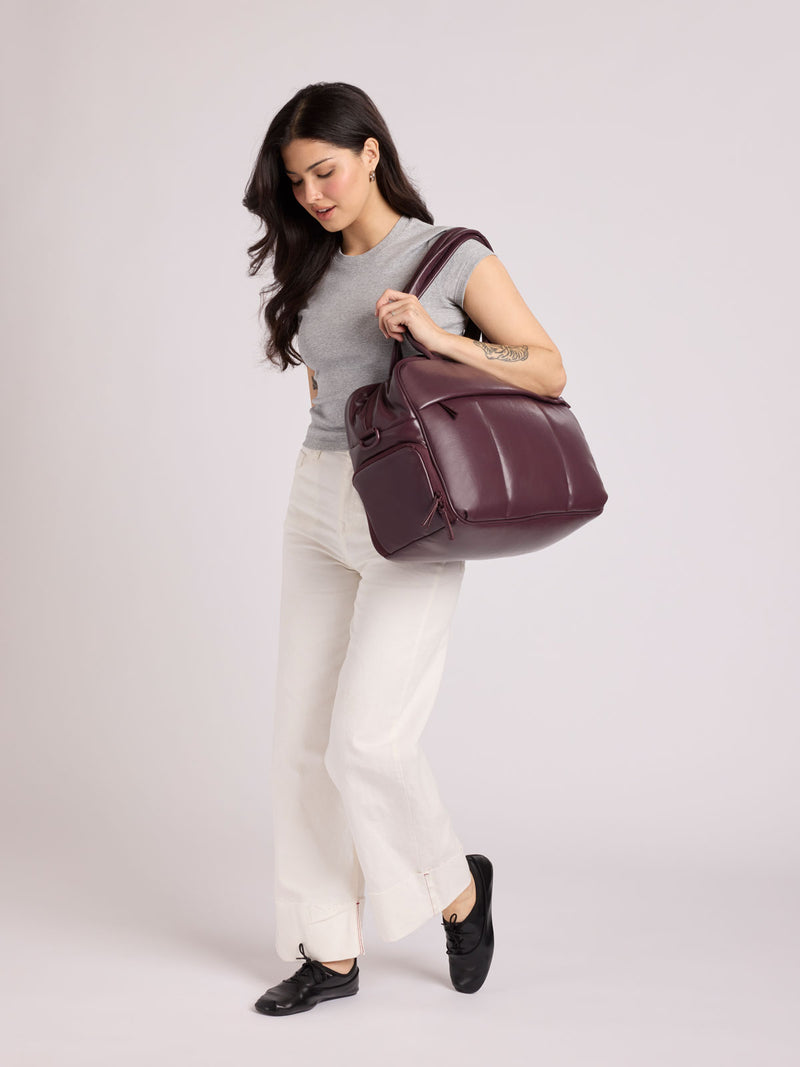 A woman wearing a gray sleeveless top, white pants, and black shoes carries a  dark burgundy CALPAK Luka Duffel in vegan leather with dual top handles and a front zippered pocket, photographed against a clean white background