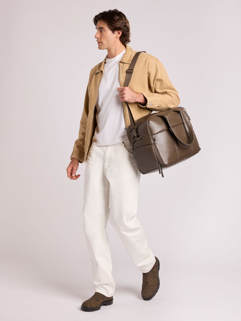 A man wearing a tan jacket, white t-shirt, white trousers, and brown boots carries a  dark brown Mushroom CALPAK Luka Duffel in vegan leather with dual top handles and a front zippered pocket, photographed against a clean white background