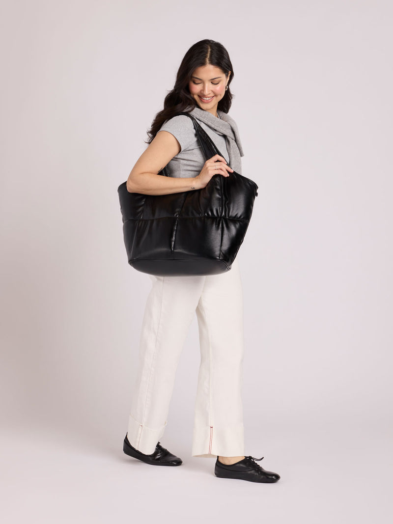 A woman modeling a CALPAK Luka Everything Tote in Black Vegan Leather, wearing a gray sleeveless top, white pants, and black shoes against a light gray studio background