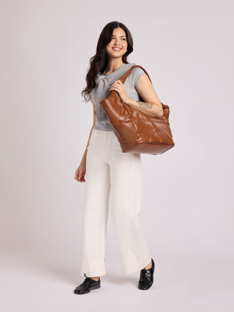 A woman modeling a CALPAK Luka Everything Tote in Honey Brown Vegan Leather over her shoulder, wearing a gray sleeveless top, white wide-leg pants, and black loafers, against a light gray studio background