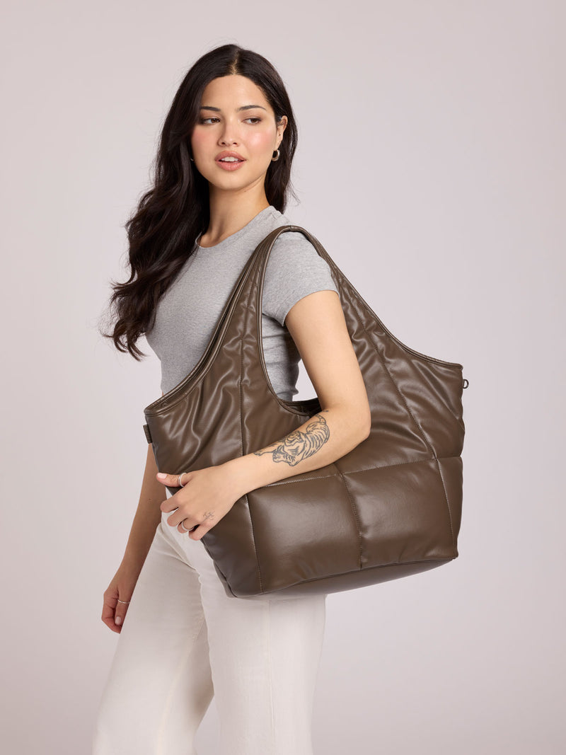 A woman wearing a gray t-shirt and white pants carries a CALPAK Luka Everything Tote in Mushroom brown Vegan Leather over her shoulder against a light gray studio background