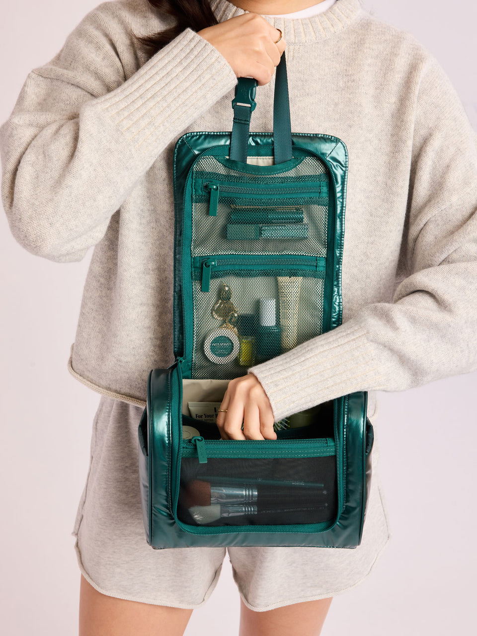 Person adding toiletries in CALPAK Luka Hanging Toiletry Bag in dark green shiny Kale; TLH2301-GLAZED-KALE