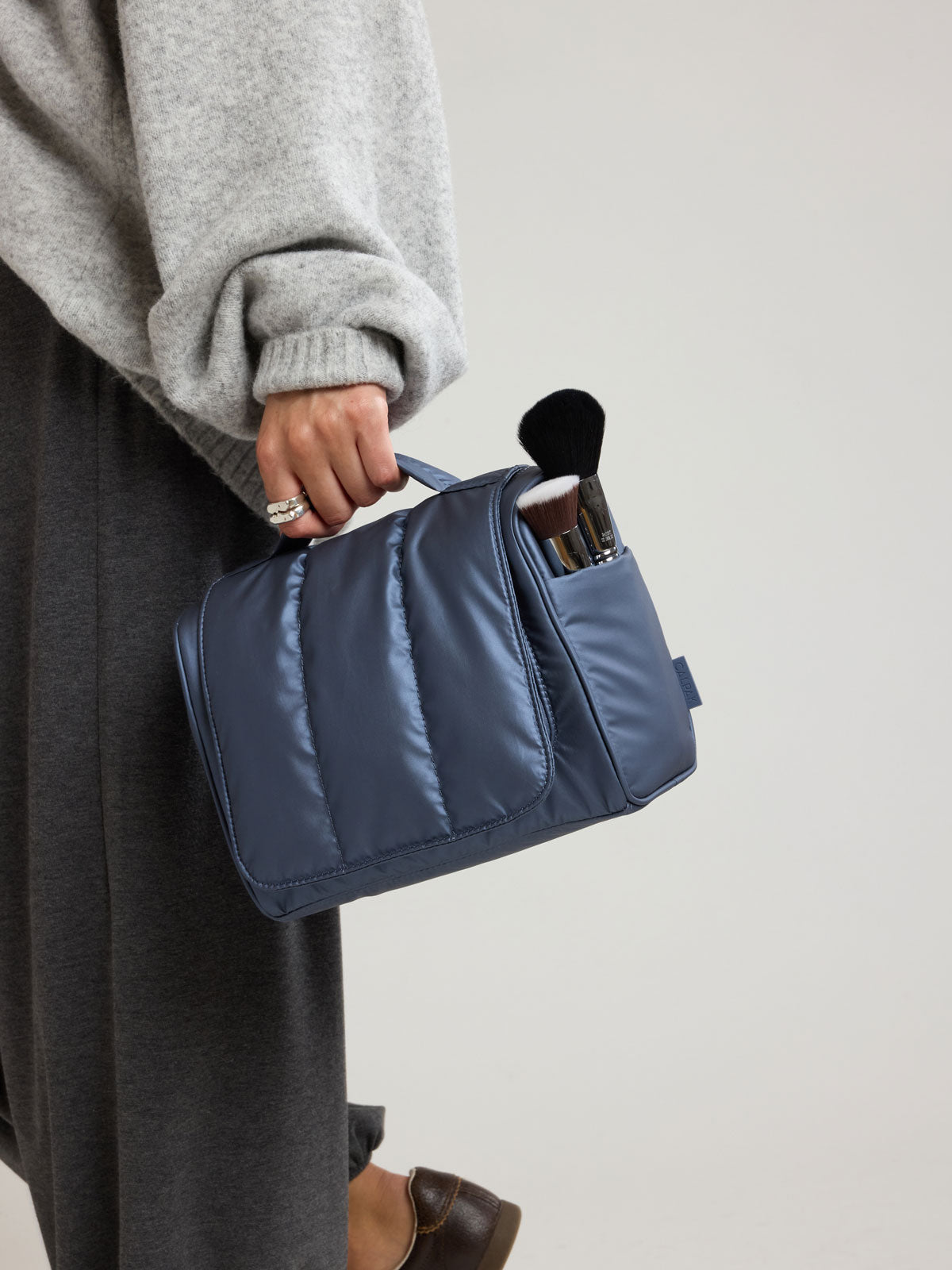 Woman carrying the CALPAK Luka Hanging Toiletry Bag in metallic dark blue material in Metallic Indigo; TLH2301-METALLIC-INDIGO