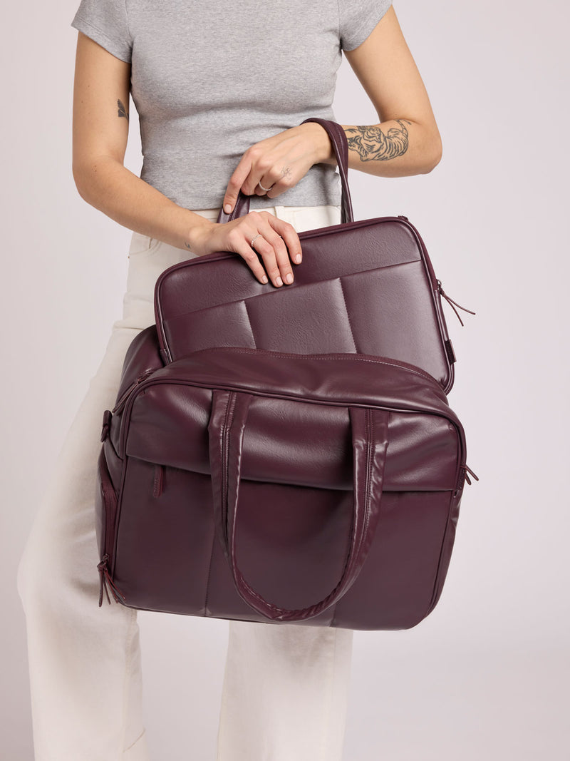 Woman wearing a grey top and white pants putting a CALPAK Luka 14in Padded Laptop Sleeve in Dark Cherry Vegan Leather into a CALPAK Luka Duffel in Dark Cherry Vegan leather against a plain background