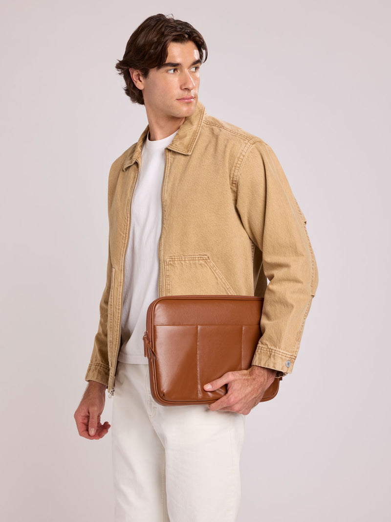 A man wearing a tan canvas utility jacket over a white t-shirt and white pants, holding a CALPAK Luka 14in Padded Laptop Sleeve in Honey Brown Vegan Leather