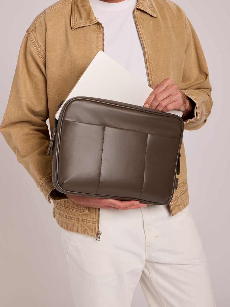 A person wearing a tan jacket and white pants holds a CALPAK Luka 14in Laptop Sleeve in Mushroom brown Vegan Leather with a white laptop inside, against a light gray studio background