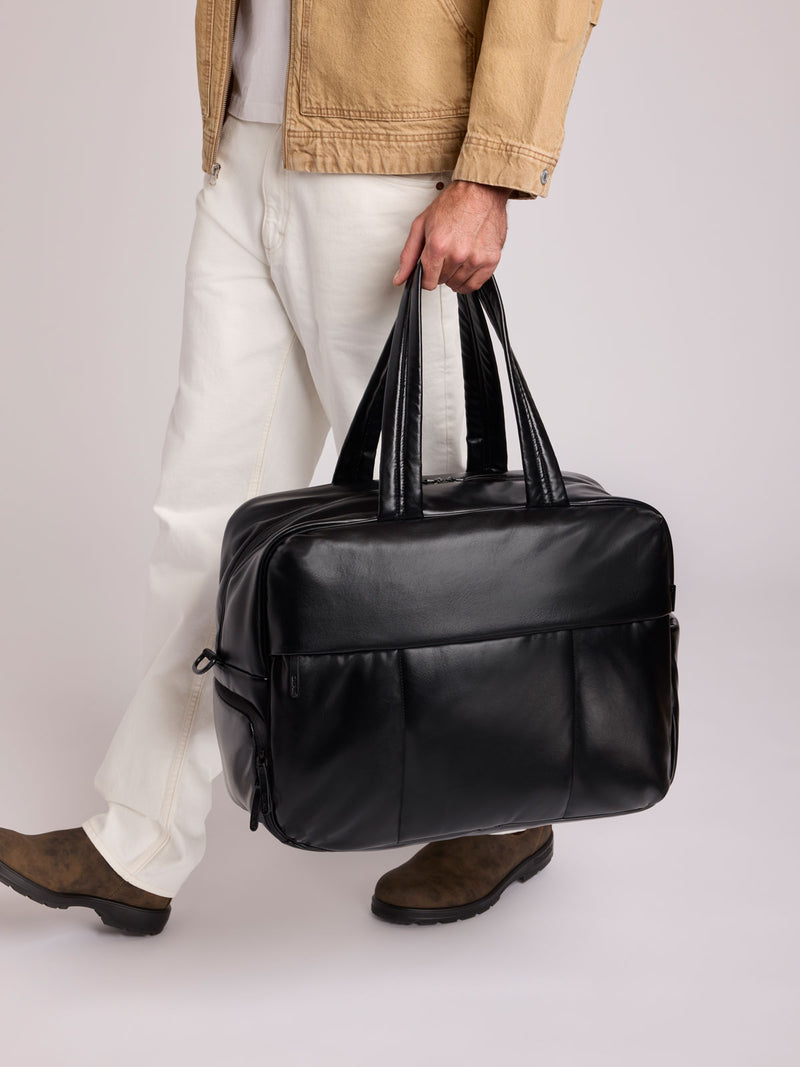 A person in a tan jacket, white pants, and brown shoes carries a Black CALPAK Luka Duffel in vegan leather with dual top handles and a front zippered pocket, photographed against a clean white background