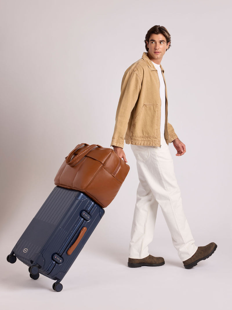 A man in a beige jacket, white shirt, and white trousers walks while pulling a CALPAK Vault Luggage suitcase with a Honey Brown CALPAK Luka Duffel in vegan leather with dual top handles and a front zippered pocket, photographed against a clean white background