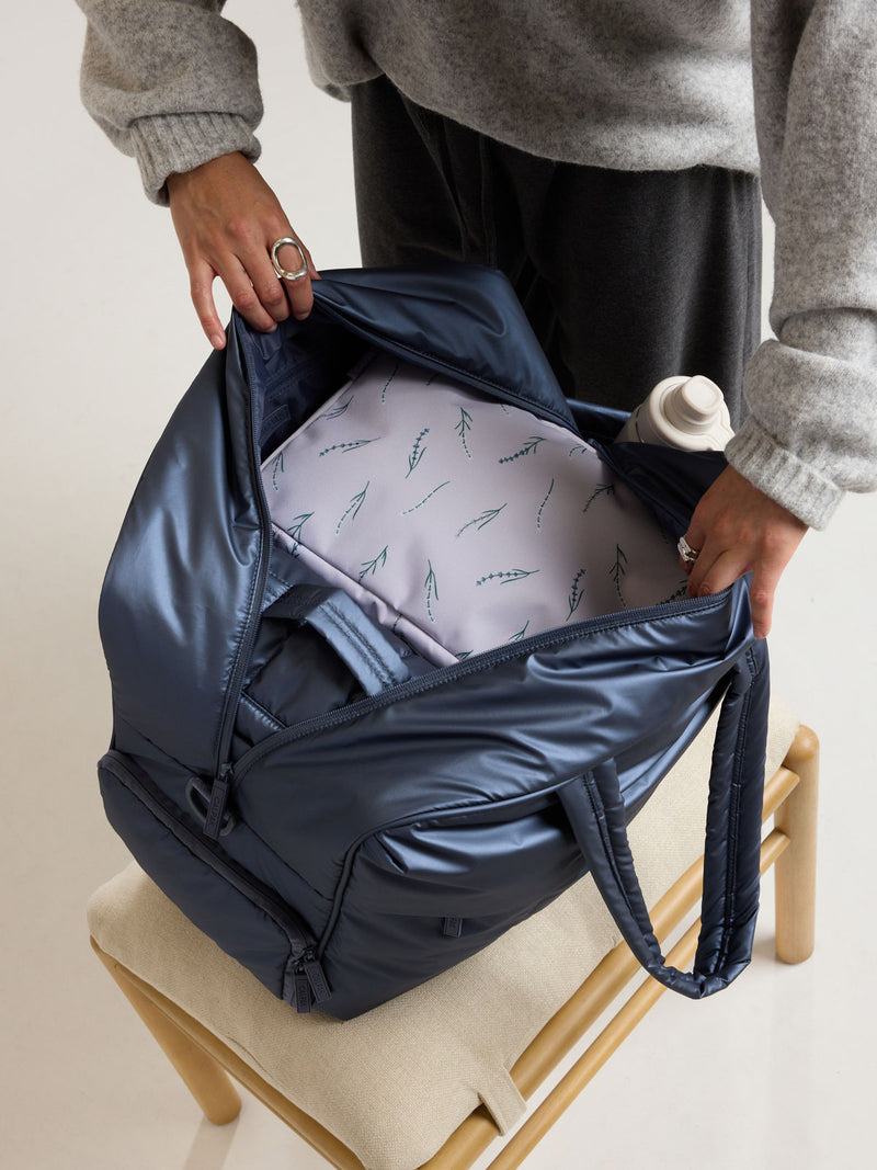 Model opening the CALPAK Luka Large Duffel in dark blue metallic material in Metallic Indigo