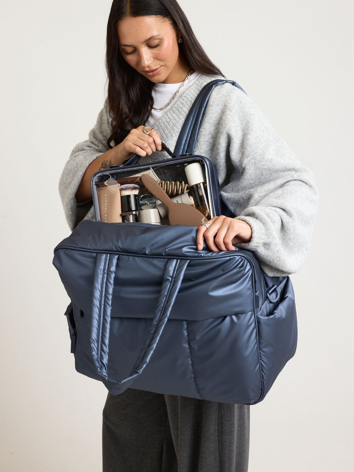 Model adding items into CALPAK Luka Large Duffel in dark blue metallic material in Metallic Indigo; DLL2201-METALLIC-INDIGO