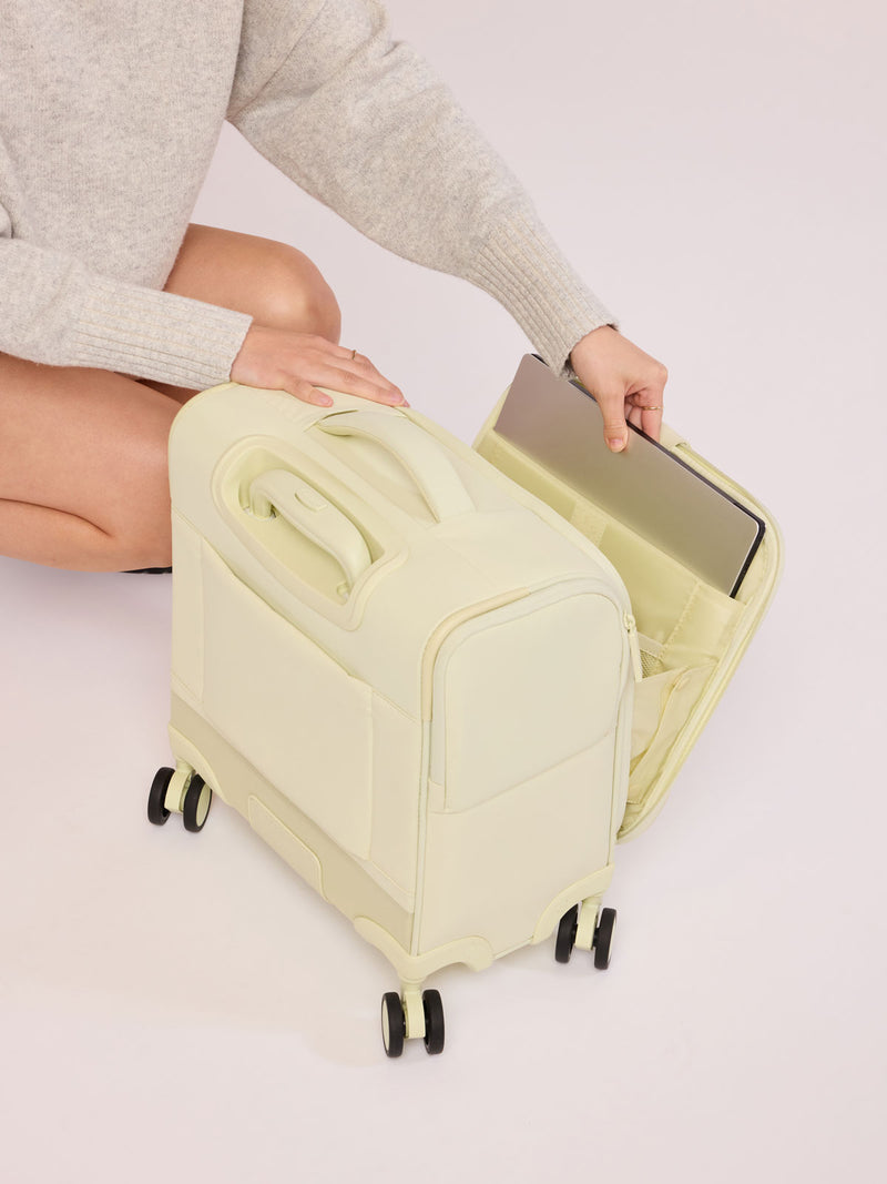 Model putting laptop in the laptop sleeve of CALPAK Luka Soft-Sided Mini Carry On Luggage in light green Celadon with spinner wheels and handle