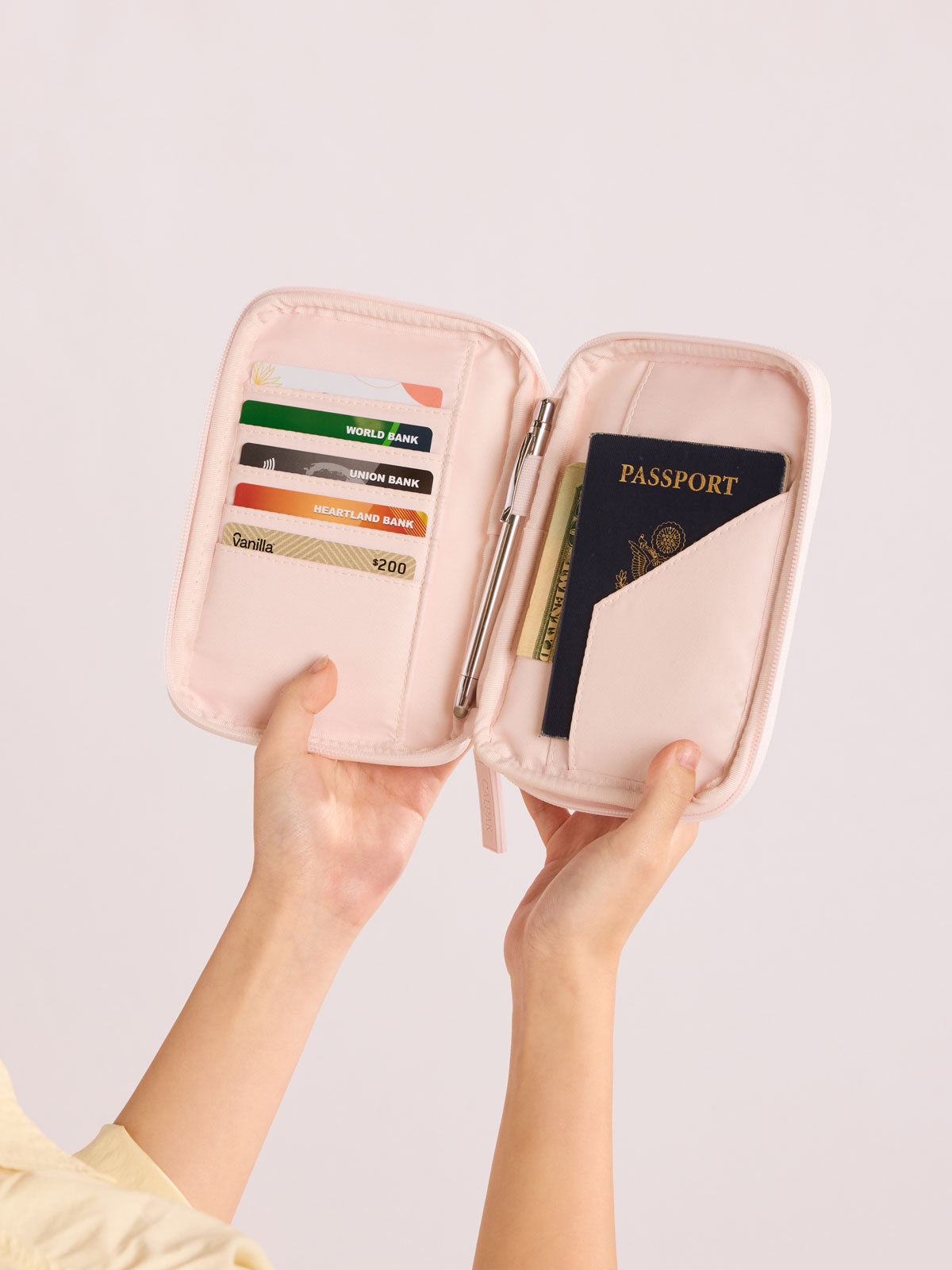 Person holding open CALPAK Luka Zip Passport Wallet in light pink shiny Pearlescent; PZW2301-PEARLESCENT